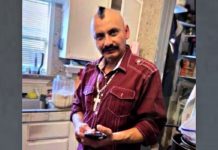 Man with a mohawk and maroon shirt in a kitchen, looking at a smartphone in his hands with cross necklace visible.
