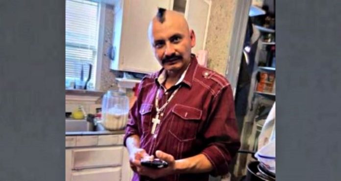 Man with a mohawk and maroon shirt in a kitchen, looking at a smartphone in his hands with cross necklace visible.
