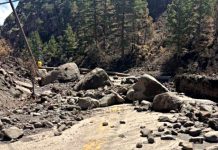 Beaver Canyon closed after severe flooding, roadway washout Rockslide blocks a dirt road in a forest, with large boulders and loose stones across the path (worker in yellow in distance).