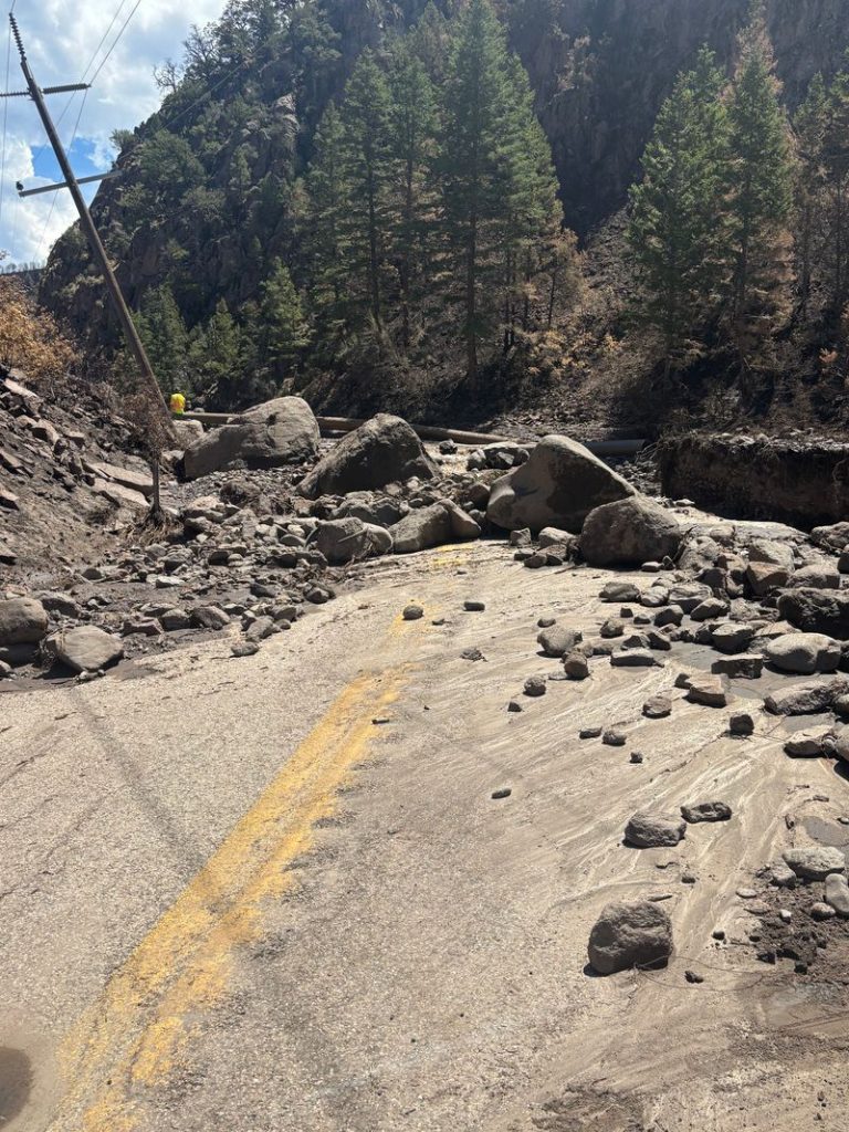 Mountain road blocked by a rockslide with large boulders and dirt a worker in a yellow jacket visible in the distance