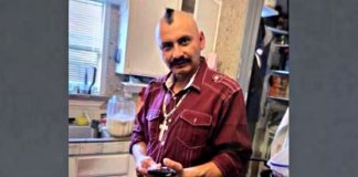 Man with a shaved head and mustache in a maroon shirt, holding a smartphone in a home kitchen background with appliances nearby.