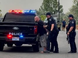 Man in custody after allegedly firing gun in Bluffdale hotel room when police responded Arrest scene: two police officers detain a barefoot man near a black police pickup with flashing red and blue lights in a parking lot.
