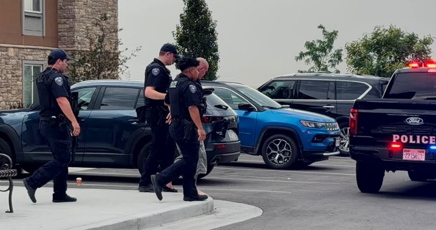 Four police officers escort a handcuffed man through a parking lot beside a gray SUV; a police truck sits nearby with lights on.