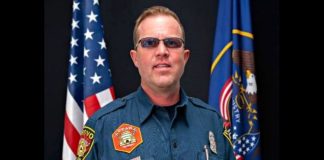 Male emergency services officer in a teal uniform with patches, posed in front of U.S. and state flags behind him.