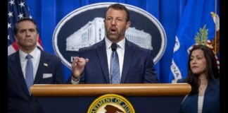 Man in a navy suit speaks at a podium with a DOJ seal, flanked by two aides during a press conference.