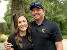 Smiling couple outdoors, woman in black top and man in a polo shirt with a bee logo posing together in a yard.