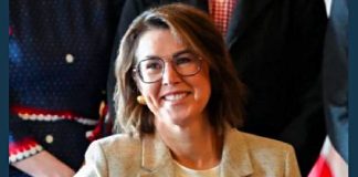 Smiling woman with short hair and glasses wearing a beige blazer at a formal event, group in background.