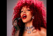Woman with bright red curly hair applying bold red lipstick, wearing diamond earrings and a jeweled choker necklace in a glam portrait.
