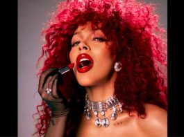 Doja Cat announces Oct. 10 concert stop at Maverik Center Woman with bright red curly hair applying bold red lipstick, wearing diamond earrings and a jeweled choker necklace in a glam portrait.