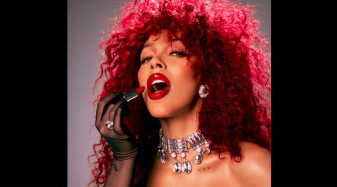 Woman with bright red curly hair applying bold red lipstick, wearing diamond earrings and a jeweled choker necklace in a glam portrait.