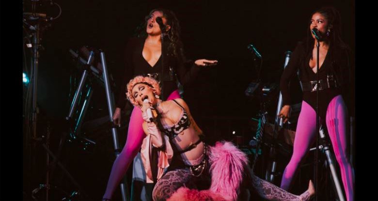 Three female performers on a dark stage; center kneels with a microphone and pink feather boa, flanked by two singers in bold outfits.