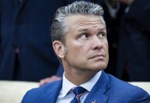Man in a blue suit and striped tie sits looking up and to the right in a formal setting, neutral expression.