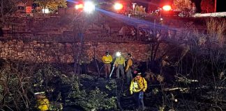 Nighttime scene of firefighters in yellow gear working among charred brush near a stone wall, with red emergency lights in the background.