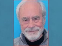 Portrait of an older man with white hair and a beard, smiling, wearing a gray sweater against a blue background.