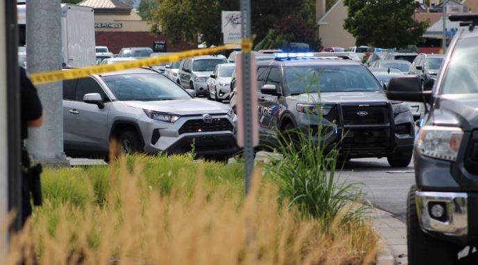 Yellow caution tape in the foreground with police patrol cars parked in a lot behind it, lights flashing on some vehicles, suggesting a scene investigation.
