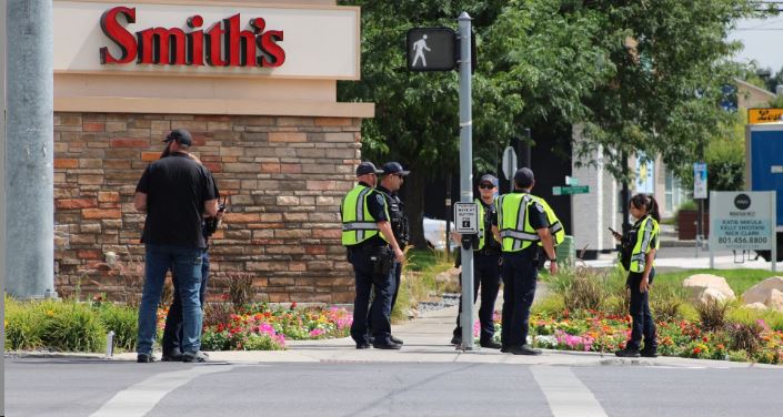 Police officers in neon vests stand at a crosswalk outside a Smith's store, with flower beds nearby.
