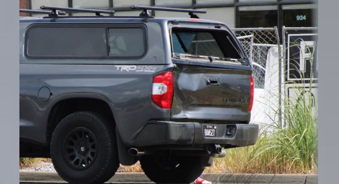 Rear three-quarter view of a gray Toyota Tundra pickup with a dented tailgate, parked near a chain-link fence and building. (informative)