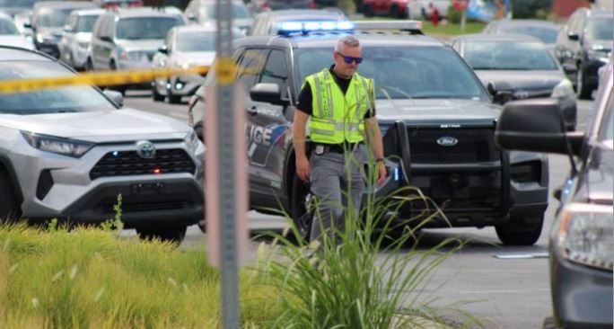 Male officer in a neon safety vest walking through a police crime-scene area with patrol cars and yellow tape in the background.