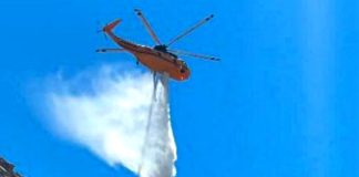 Orange firefighting helicopter releasing a large white water load over a rocky cliff against a clear blue sky