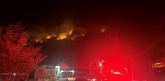 Nighttime view of a residential neighborhood with a hillside fire and flames above, emergency lights glow red on the scene behind the houses.