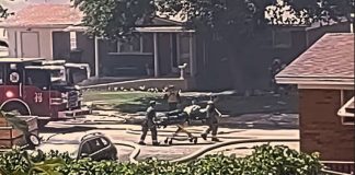 Firefighters and bystanders push a stretcher along a suburban street with a fire truck nearby.