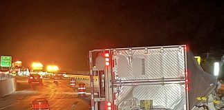 Nighttime highway construction scene with a large silver trailer, orange traffic barrels, and flashing lights blocking the road ahead.