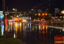Water main break closes westbound North Temple at Redwood Road in Salt Lake City Nighttime city street flooded with reflections on the wet road, orange construction barrel in the foreground and distant city lights and signs (Denny’s, Chevron).
