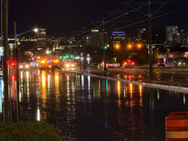 Water main break closes westbound North Temple at Redwood Road in Salt Lake City Nighttime city street flooded with reflections on the wet road, orange construction barrel in the foreground and distant city lights and signs (Denny’s, Chevron).