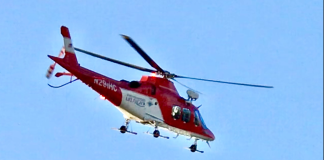 Red-and-white rescue helicopter in flight against a clear blue sky, landing gear visible beneath the fuselage.