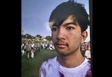 Close-up of a young man with dark hair and light stubble at an outdoor park, with people and green grass in the background under a blue sky.