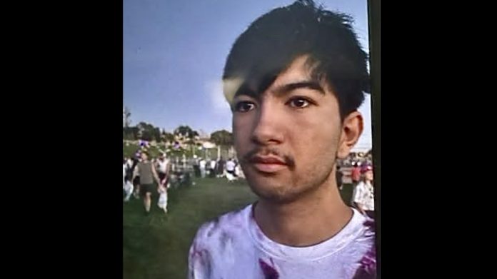 Missing Weber County 18-yea-old Close-up of a young man with dark hair and light stubble at an outdoor park, with people and green grass in the background under a blue sky.