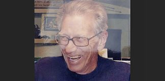 Older man with glasses smiling and laughing, in a dark shirt.