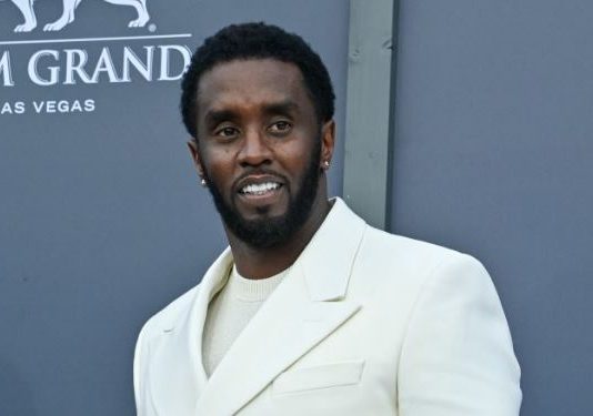 Sean ‘Diddy’ Combs in solitary confinement after prison fight Man in a cream-colored suit posing on a blue-gray backdrop at MGM Grand Las Vegas, smiling for the camera on a red carpet event.