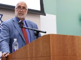 SLCo DA declines to file charges against Sandy PD officers after fatal shooting A bearded man in a blue suit and red tie speaks at a wooden podium with a microphone, a water bottle nearby, and a large emblem backdrop partially visible behind him.