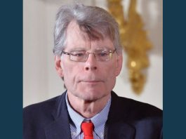 Stephen King responds to Utah ban of several classic novellas Older man with gray hair and glasses wearing a dark suit, light blue striped shirt, and red tie, looking forward with a neutral expression; blurred gold decor in the background.