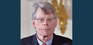 Stephen King responds to Utah ban of several classic novellas Older man with gray hair and glasses wearing a dark suit, light blue striped shirt, and red tie, looking forward with a neutral expression; blurred gold decor in the background.