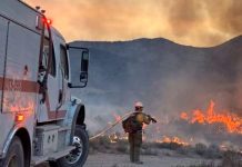 Stookey Fire hits 10.3K acres, 20% contained Firefighter in turnout gear spraying water from a hose as a fire truck battles a brush fire in a desert at sunset.