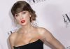Taylor Swift marries Travis Kelce in NYC; Adam Sandler officiates Woman with brown hair in an updo, bold red lipstick, gold drop earrings, wearing a strapless black dress at a formal event.