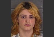 Mugshot photo of a young person with shoulder-length orange-blonde hair and a neutral expression against a gray wall/background.