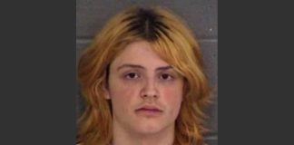 Mugshot photo of a young person with shoulder-length orange-blonde hair and a neutral expression against a gray wall/background.