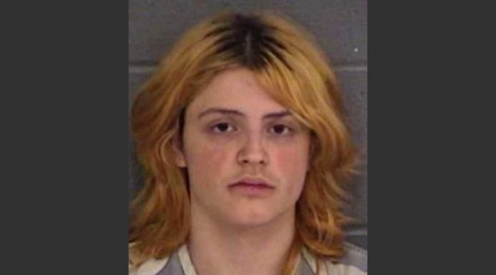 Mugshot photo of a young person with shoulder-length orange-blonde hair and a neutral expression against a gray wall/background.