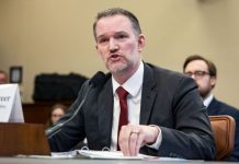 Man in a dark suit and red tie testifies at a formal hearing, speaking into a microphone with papers on the desk.