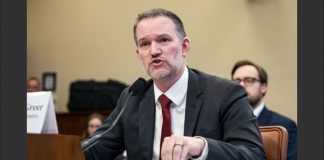Man in a dark suit and red tie testifies at a formal hearing, speaking into a microphone with papers on the desk.