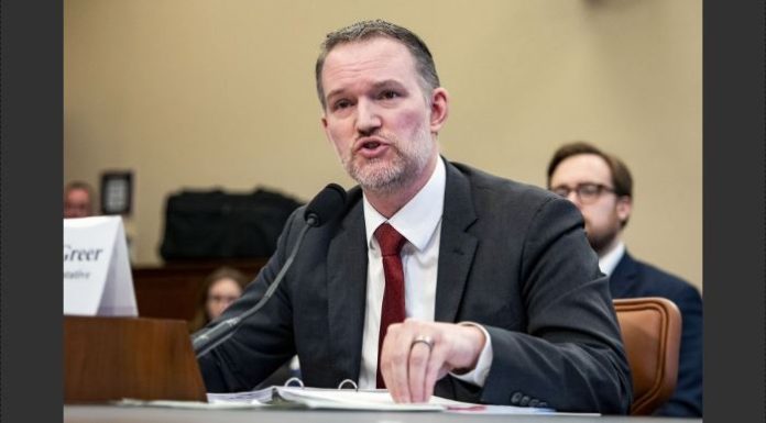 Man in a dark suit and red tie testifies at a formal hearing, speaking into a microphone with papers on the desk.