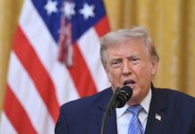 Man in a suit speaks into a microphone with an American flag and gold curtains behind him.