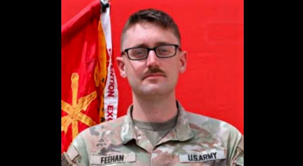 US Army soldier in camouflage uniform with name tag Feehan black framed glasses and mustache posed in front of a red backdrop and flag