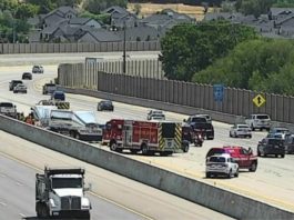 Emergency responders on a multilane highway incident with fire trucks and police cars, concrete barriers, and houses in the distance on a residential hillside. A signaled lane shows activity and partial closures behind the barrier.