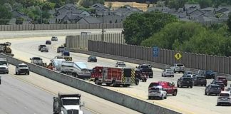 Emergency responders on a multilane highway incident with fire trucks and police cars, concrete barriers, and houses in the distance on a residential hillside. A signaled lane shows activity and partial closures behind the barrier.