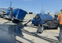 Highway crash scene with a crushed blue car and a blue semi-trailer, fluids spilled on the road, and first responders in orange vests nearby.