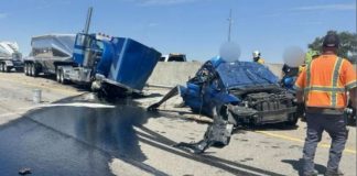 Highway crash scene with a crushed blue car and a blue semi-trailer, fluids spilled on the road, and first responders in orange vests nearby.